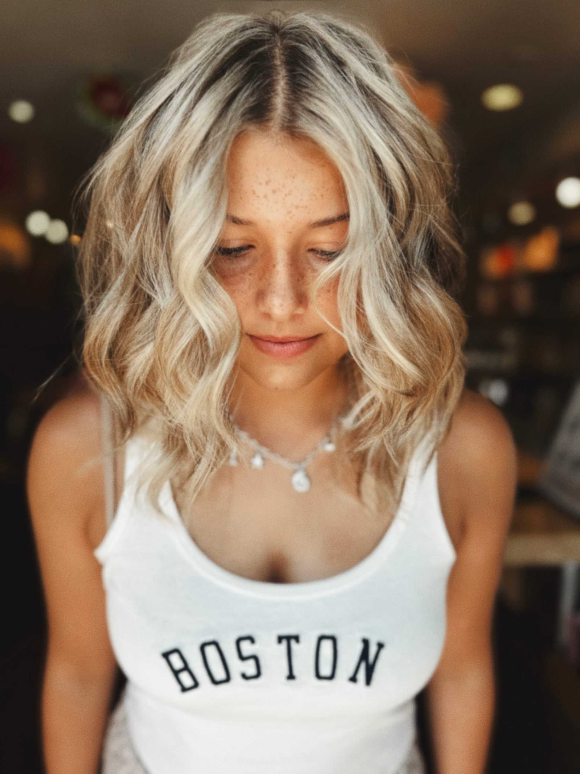 Blonde woman with wavy hair in a "Boston" tank top, looking down in a softly lit setting.