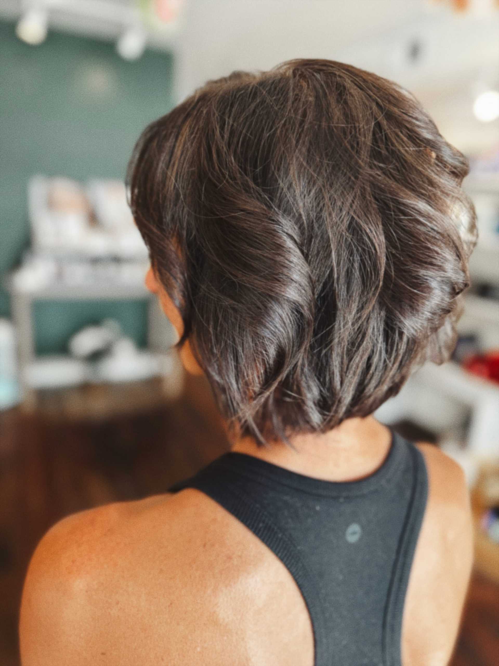 Short-haired person in a black top is standing, viewed from behind in a salon setting.